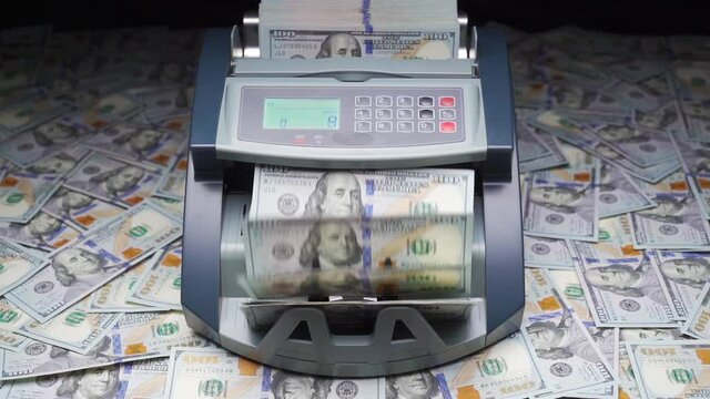 Currency counting machine stands on heap of hundred dollars. Cash bill counter money machine counts 100 dollar bills or USD banknotes. Automatic mechanism for bank financial operations. Slow motion
