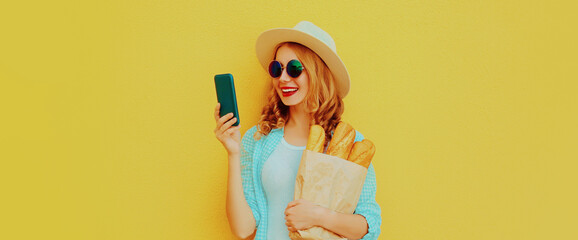 Portrait of happy smiling woman with phone holding grocery shopping paper bag with long white bread baguette on a yellow background