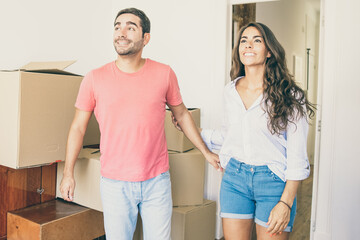 Obraz premium Happy excited young couple looking over their new apartment with carton boxes and furniture, smiling and talking. Medium shot. New home concept