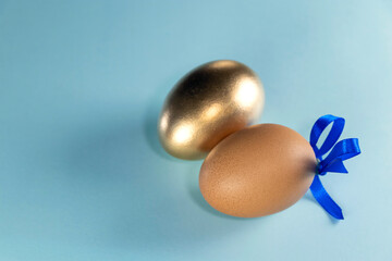 Brown egg with blue ribbon and golden egg. Happy Easter concept. Two eggs with ribbon on a pastel blue background