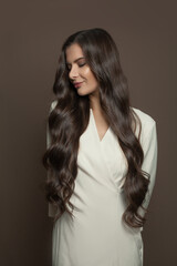 Nice woman with long dark hairstyle wearing white dress standing on brown background