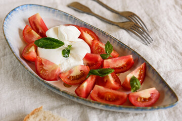 Traditional Italian salad with Burrata cheese, tomatoes and fresh basil. Mozzarella burrata salad with basil leaves and tomatoes on plate on beige linen tablecloth