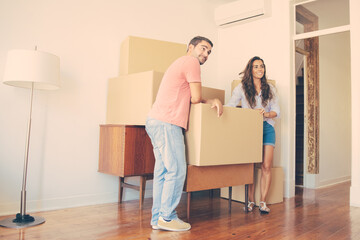 Fototapeta na wymiar Positive young couple looking over their new apartment, while standing and leaning on cardboard boxes and furniture indoors. Full length. New home or property buying concept
