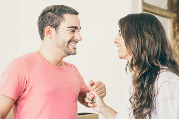 Happy dating girl and guy moving into new flat, standing near carton boxes, holding hands, talking and laughing. Medium shot. New home concept