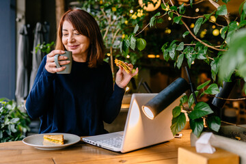 Charming female freelancer in trendy clothing working on laptop during morning breakfast at cozy cafe. Caucasian middle age woman with brown hair eating toast with cheese, top view.
