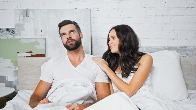 cheerful woman touching shoulder of bored boyfriend while talking to him in bed