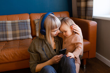 Young woman with headphones and baby at home. Mother with one year old child listening to music at home. Woman with phone, headphones and child sitting on the floor at home.