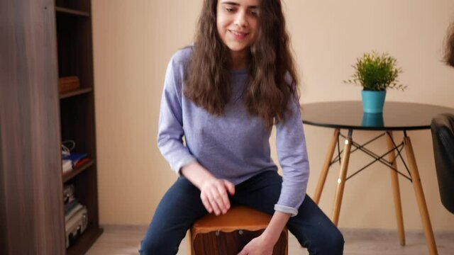 Cute teenager girl play the cajob sitting on box drum at home during rehearsal with her sister in a room