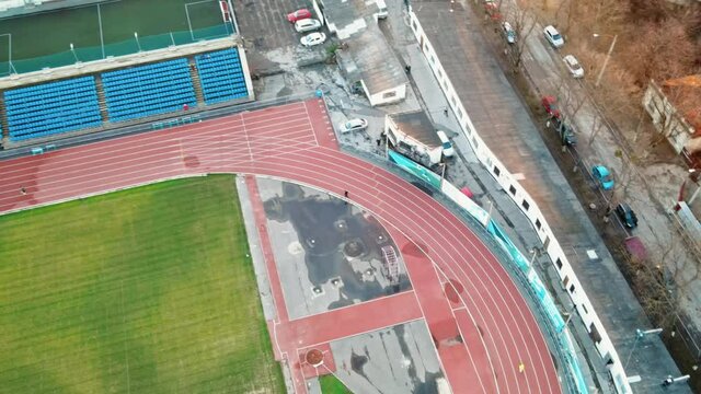 Aerial drone view of a stadium in Chisinau. Green field, running tracks with a man, tribunes, buildings nearby, roads with moving cars and bare trees. Moldova