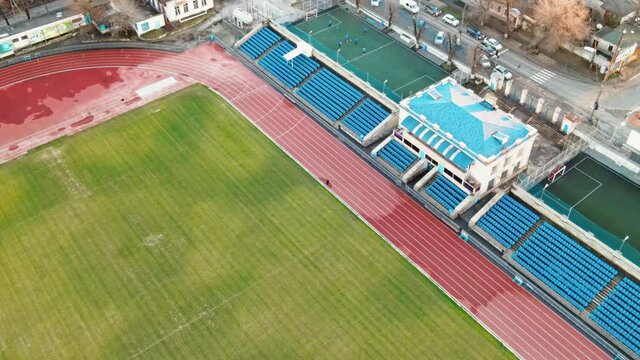 Aerial drone view of a stadium in Chisinau. Green fields, running tracks with a man, tribunes, buildings nearby, roads with moving cars and bare trees. Moldova