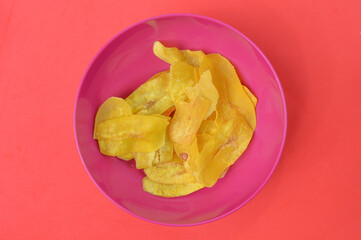 Top view of banana slice chips in a plastic bowl over red background.