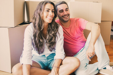 Joyful Latin couple in love enjoying moving into new apartment, sitting on floor near stacks of carton boxes, looking away and laughing. New home or relocation concept