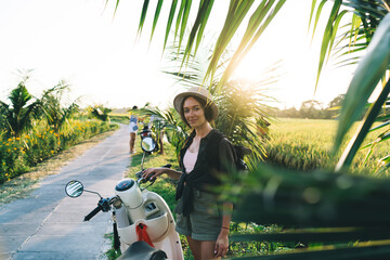 Portrait of beautiful female traveller in trendy straw hat looking at camera during tropical vacations, half length of hipster girl posing near palm trees and retro scooter enjoying summer wanderlust © BullRun