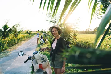 Fototapeten Bali Portrait of beautiful female traveller in trendy straw hat looking at camera during tropical vacations, half length of hipster girl posing near palm trees and retro scooter enjoying summer wanderlust  © BullRun