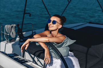 Portrait of smiling happy girl in striped dress and sunglasses sitting at the steering wheel of her own expensive yacht and looking at the road. Laxury concept.