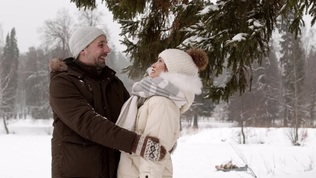 Medium of cheerful Caucasian man hugging, talking to young Asian woman under pine tree in snowy winter park. Couple wearing warm clothes, getting interrupted by child throwing snow, separating them 