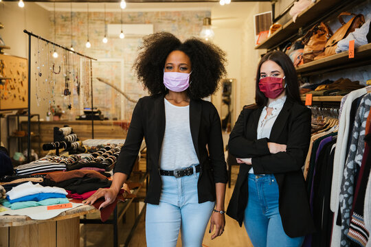 Portrait of two women owners of the clothes shop at the entrance to welcome customers during the Coronavirus Covid-19 pandemic wearing protective face masks - Millennial initiate a start-up business