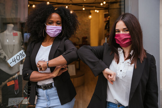 Portrait two saleswomen in front of clothing store greeting each other giving strength touching elbows wearing protective face masks in Coronavirus Covid-19 pandemic Millennial sales assistant smile