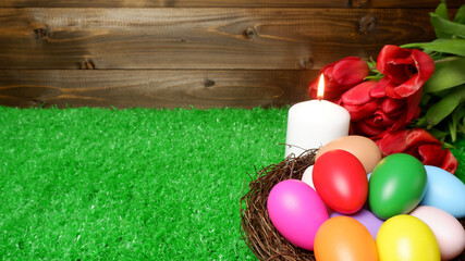 Burning white wax candle, multi colored easter eggs in birds nest and bouquet of red tulip flowers on green grass lawn and wooden panks background closeup view selective focus with copy space