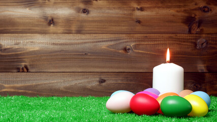 Burning white wax candle surrounded by multi colored easter eggs on green grass lawn and wooden panks background closeup view selective focus with copy space. Easter holiday banner, card, poster
