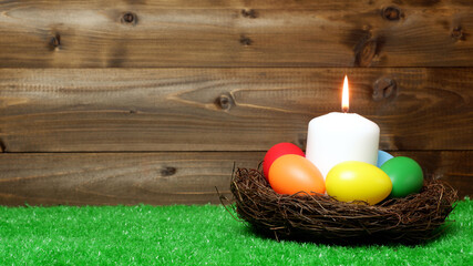 Burning white wax candle in birds nest surrounded by multi colored easter eggs on green grass lawn and wooden panks background closeup view selective focus with copy space