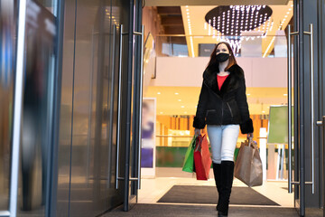 Beautiful woman in protective mask with shopping bags walks out mall. Stylish shopper girl