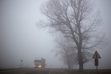 The truck is driving on a foggy road with bare trees, dangerous weather conditions. Driving in poor visibility