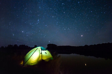 Illuminated tent near the river and beautiful sky full of stars. Night travelling landscape © Vitalii