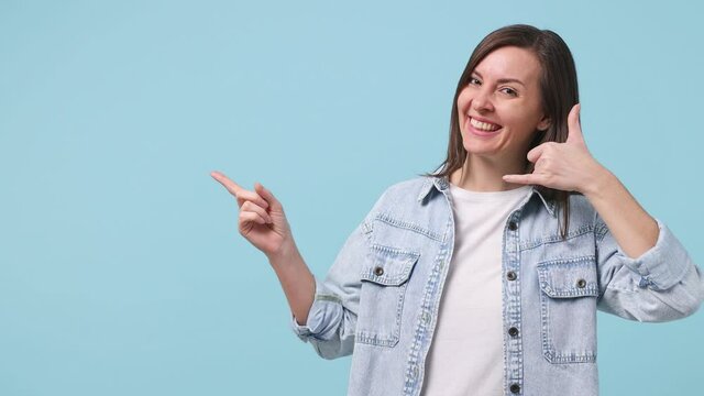 Smiling young woman 30s years old in denim jacket white t-shirt doing phone gesture like says call me back pointing aside on workspace copy space mock up area isolated on pastel blue background studio