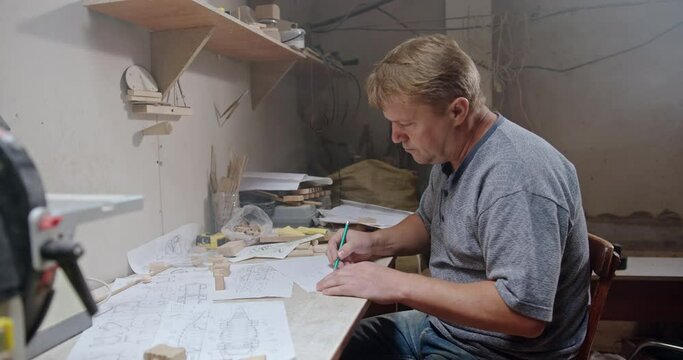 Artisan man drawing sketch of wooden detail in workshop. Side view of focused middle aged carpenter creating draft of wooden detail while sitting at table in professional workshop