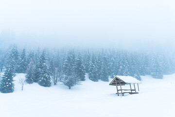 Solitary wooden shelter against a backdrop of mountains and forest during heavy snowfall and fog.