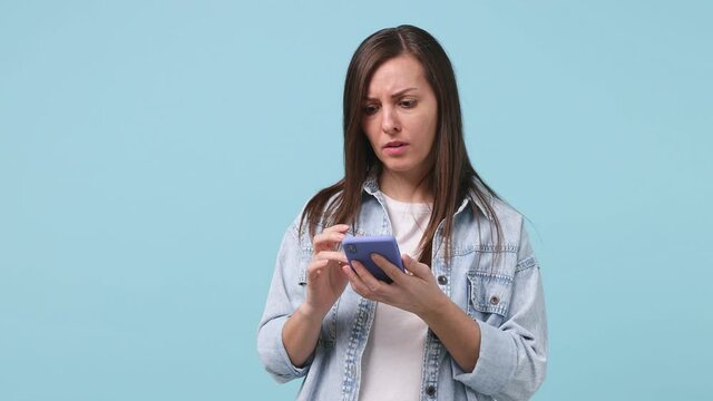 Dissatisfied irritated young woman 30s years old in denim jacket white t-shirt use mobile cell phone typing browsing swear showing thumb down dislike gesture isolated on pastel blue background studio
