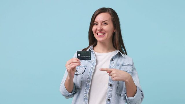 Smiling long hair brunette young woman 30s years old in denim jacket white t-shirt pointing index finger on mockup plastic credit bank card showing thumbs up isolated on pastel blue background studio