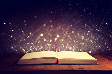 Magical image of open antique book over wooden table with glitter overlay