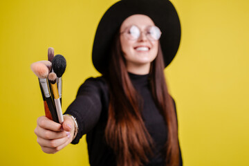 Girl in black clothes in a big black hat and glasses standing on a yellow background holding out makeup brushes forward