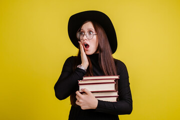 Beautiful girl in black clothes in a big black hat and glasses, standing on a yellow background with a stack of books, thinking with horror about exams