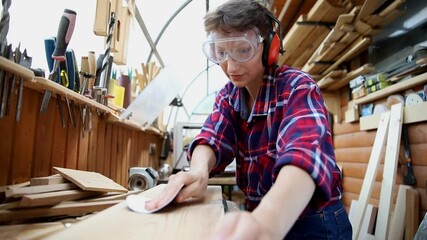 Young attractive 30-40 professional female carpenter looking grinding raw wood with sandpaper in carpentry diy workshop. Feminine women equality concept. Adult girl with male hobby at workbench