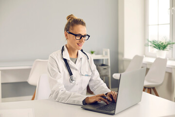 Friendly young doctor sitting at desk, using laptop computer, doing medical Internet research, entering electronic health records into database or giving online consultation to patient on eHealth chat