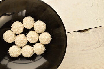 Three delicious sweets with coconut flakes on a black ceramic plate, on a wooden table, close-up, top view.