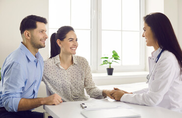 Smiling couple and family doctor sitting at desk in office, looking at each other and holding hands. Visit to hospital, planning pregnancy, happy patients, satisfied clients, trust and respect concept