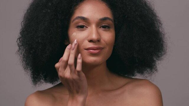 smiling african american woman applying face foundation isolated on grey