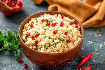 Cooked couscous with parsley and pomegranate in a ceramic bowl on a dark background close-up. Maghreb cuisine, vegan food.