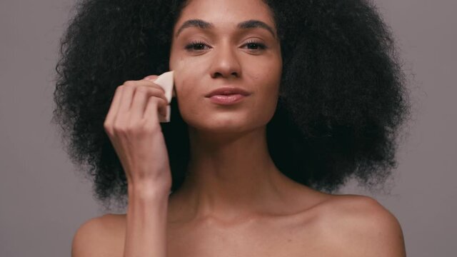 african american woman applying makeup foundation with sponge isolated on grey