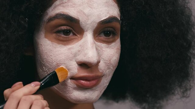 african american woman applying clay mask with cosmetic brush isolated on grey