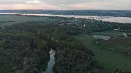 Aerial view of a green forest with a small river. Automobile bridge over the Dnieper river in the background. There is a city on one bank of the river and summer cottages on the other bank.