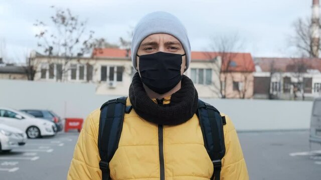 Food courier with backpack and black medical mask on parking, watching into camera. Food delivery service
