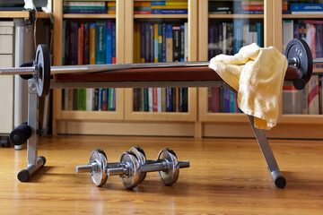 Home gym equipment: iron dumbbells beneath a weight bench with a long barbell and a towel in home office. - Powered by Adobe