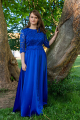 Portrait of an attractive girl standing in nature after high school graduation. Beautiful smiling female model in elegant dress posing at summer park looking at camera motion shot.