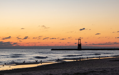 orange and yellow early morning sky. sunrise over the beach. landscape of waves crashing on the beach shoreline at sunrise. seagulls by the lakefront at dawn. silhouette of beach tower during sunrise