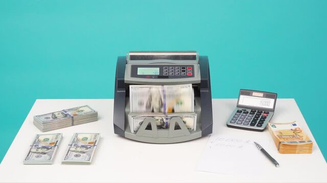 Man puts money 100 dollar bills or USD banknotes in counting money machine. Finance concept. Currency counter machine stands on white table in office isolated on blue. Bank financial operations
